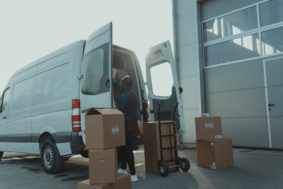 A professional mover loading a white cargo van parked outside a modern building with large glass windows during daylight. The mover, dressed in dark clothing and wearing a cap, is using a hand truck t