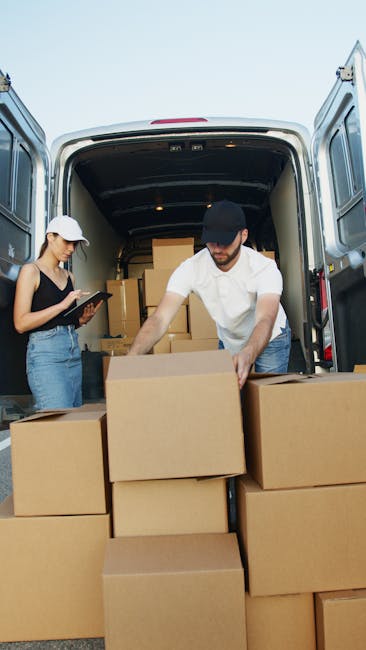 A professional mover loading a white cargo van parked outside a modern building with large glass windows during daylight. The mover, dressed in dark clothing and wearing a cap, is using a hand truck t