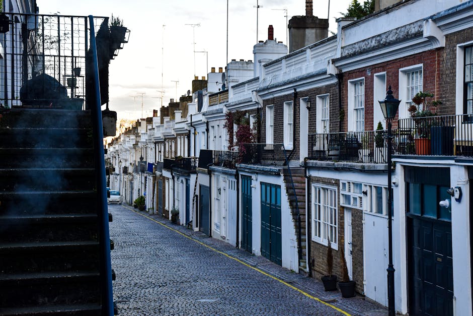 The image shows a cobbled street in Walthamstow Village lined with narrow terraced houses, each with distinct entry doors, small front gardens or steps, and wrought iron railings. The buildings are predominantly brick with some painted white or in natural brick tones, featuring sash windows, chimneys, and decorative detailing along the rooftops. Potted plants and flower boxes are visible on some front balconies, while classic street lamps illuminate the scene. The scene is captured during daylight hours with a clear sky, and the street appears quiet with no visible pedestrians or vehicles. Along one side of the street, a black staircase with open metal steps and a railing is partially visible, possibly leading to an upper level or side entrance. The overall setting reflects a typical residential area suited for home relocation or moving services targeting narrow, terraced homes in E17, as often managed by companies like [COMPANY_NAME], focusing on packing and furniture transport in confined urban spaces.