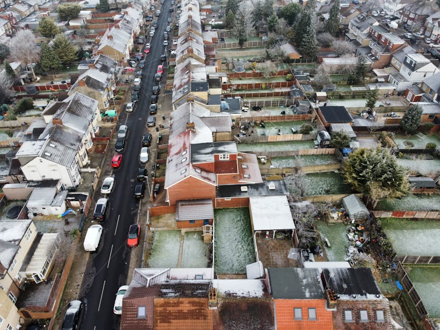 An aerial view of a residential street in Walthamstow Village covered with light snow, showing terraced houses, parked cars along the road, and back gardens enclosed by wooden fences. The gardens contain various objects such as garden furniture, sheds, and potted plants. The street features a single lane with cars parked on both sides, and the houses have pitched roofs with some snow accumulation. This scene illustrates a typical neighbourhood where local house removals or furniture transport services by [COMPANY_NAME] might be coordinated for home relocation or moving projects within the E17 area, highlighting the typical environment for loading and unloading activities in narrow terraced streets during winter.