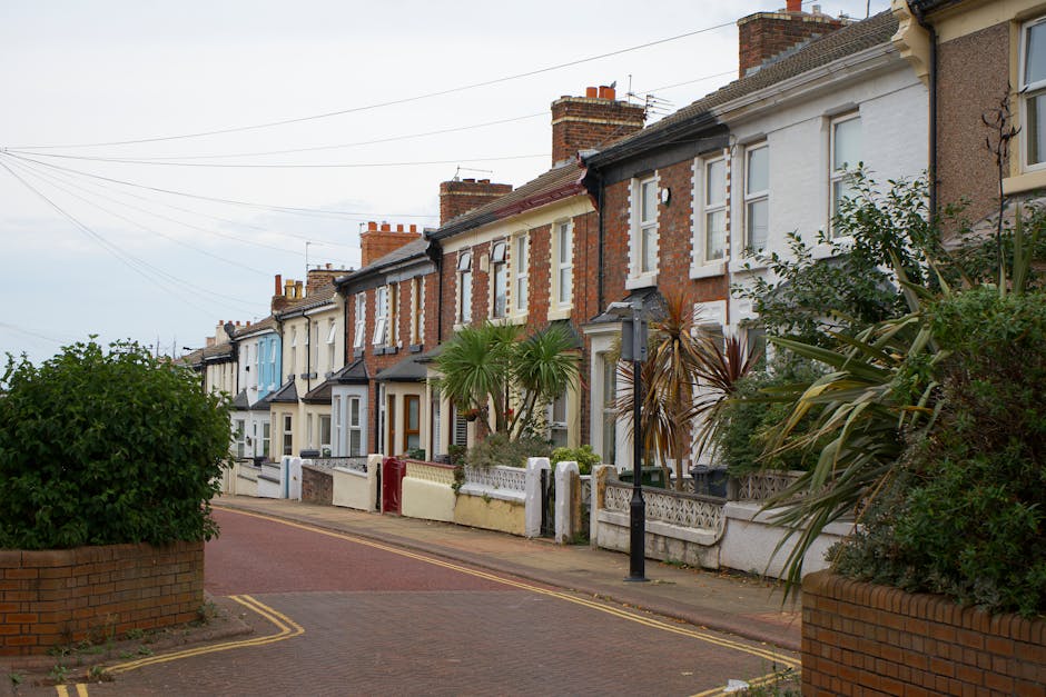 The image shows a cobbled street in Walthamstow Village lined with narrow terraced houses, each with distinct entry doors, small front gardens or steps, and wrought iron railings. The buildings are predominantly brick with some painted white or in natural brick tones, featuring sash windows, chimneys, and decorative detailing along the rooftops. Potted plants and flower boxes are visible on some front balconies, while classic street lamps illuminate the scene. The scene is captured during daylight hours with a clear sky, and the street appears quiet with no visible pedestrians or vehicles. Along one side of the street, a black staircase with open metal steps and a railing is partially visible, possibly leading to an upper level or side entrance. The overall setting reflects a typical residential area suited for home relocation or moving services targeting narrow, terraced homes in E17, as often managed by companies like [COMPANY_NAME], focusing on packing and furniture transport in confined urban spaces.
